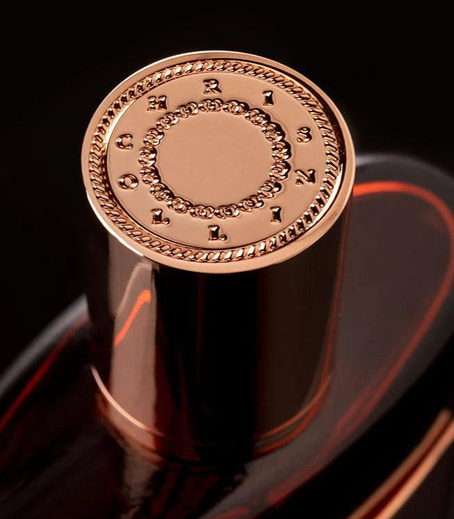 Close-up of a luxury perfume bottle featuring a highly polished rose gold cap with engraved Roman numerals and a rope-textured border, set against a dark black background.
